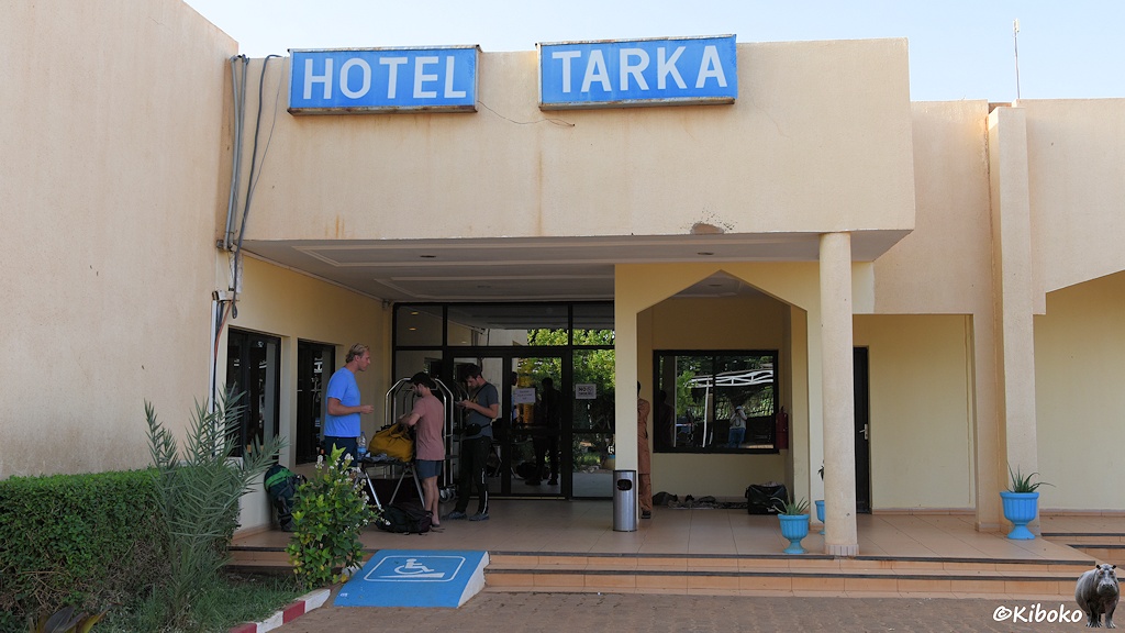 Das Bild zeigt den Eingangsbereich eines Hotels mit einer Glastür. Darüber stehen zwei blaue Schilder mit weißer Schrift: Hotel und Tarka. Drei junge Männer mit Gepäck warten vor der Tür. Im Vordergrund sind zwei Stufen und eine blau lackiete Rampe mit Rollstuhlsymbol.