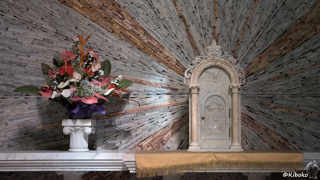 Das Bild zeigt ein Blumengesteck in einer weißen Vase auf einem Marmor Sideboard. Darauf steht eine Steintafel. Der Hintergrund ist aus graußen und rotbraunem Stein, die einen Stern hinter der Steintafel bilden.