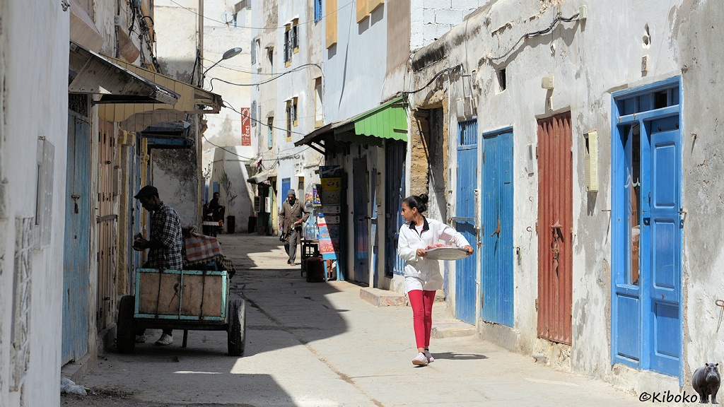Das Bild zeigt eine Gasse mit Betonoberfläche, weißen Hauswänden, überwiegend blauen Türen. Eine Frau mit roter Hose und weißer Jacke kommt mit einer Schüssel entgegen und schaut dabei zu seite. Sie beobachtet einen Mann mit Handwagen, der links eine Tür aufschließt.