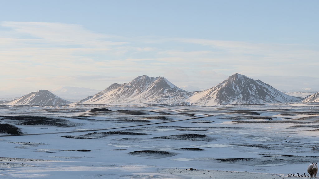 Eine Straße führt über eine schneebedeckte Ebene aus der dunkle Schotterhügel ragen. Im Hintergrund sind schroffe Berge.