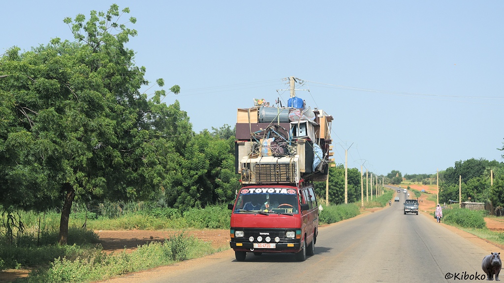 Das Bild zeigt einen roten Toyota Kleinbuss auf einer Teerstraße. Die Ladung auf dem Dach ist höher als das Fahrzeug. Über der Fahrerkabine liegt ein großer Kühlschrank. Darüber sind Möbel die kunstvoll mit Stricken gesichert sind.