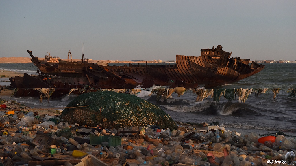 Das Bild zeigt die rostfarbende Reste vom Stahlrumpf eines Fischtrawlers, der mit der Spitze an einen völlig vermüllten Strand liegt. Plastikflaschen bedecken den Strand fast komplett. Ein Haufen Plastikmüll ist durch einen Netz gesichert. Eine Leine mit Plastikfetzen geht quer durch das Bild.
