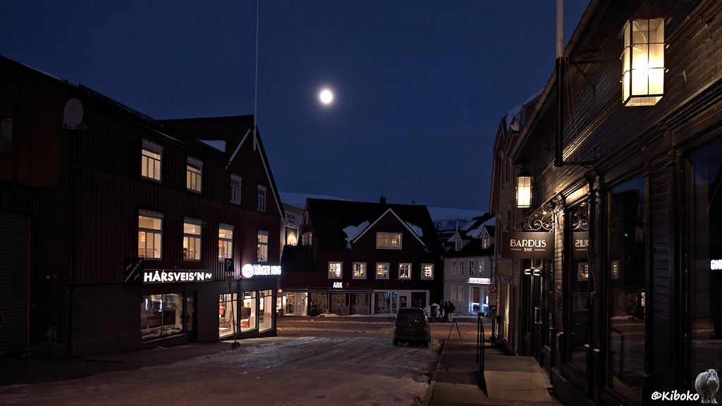 Das Bild zeigt eine Straße mit dunkelbraunen Holzhäusern. Fenster, Schaufenster und Laternen sind eingeschaltet. Darüber leuchtet der Vollmond an einem dunkelblauen Himmel.