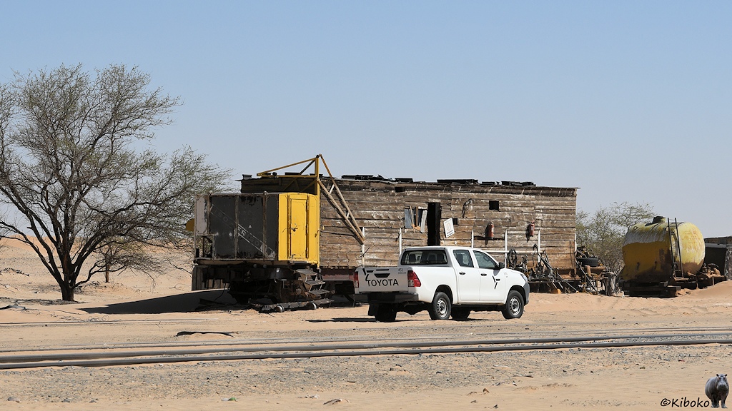 Das Bild zeigt einen Flachwagen auf dem aus dunklen Schwellen ein würfelförmiges Blockhaus errichtet wurde. Die Eingangstür in der Mitte steht offen. Am vorderen Ende des Wagens ist eine große, gelbe Blechkiste montiert. Vor dem Wagen steht ein weißer Pick-Up mit Doppelkabine.
