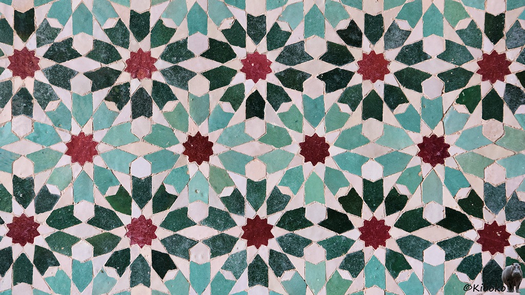 Das Bild ein Wandmosaik mit versetzten Blumen. Die Blumen bestehen aus einem roten 10-zackigen Stern mit 10 pfeilförmigen Steinchen als Blütenblätter. Die Blumenreihen haben abwechselnd dunkelgrüne und hellgrüne Blütenblätter.