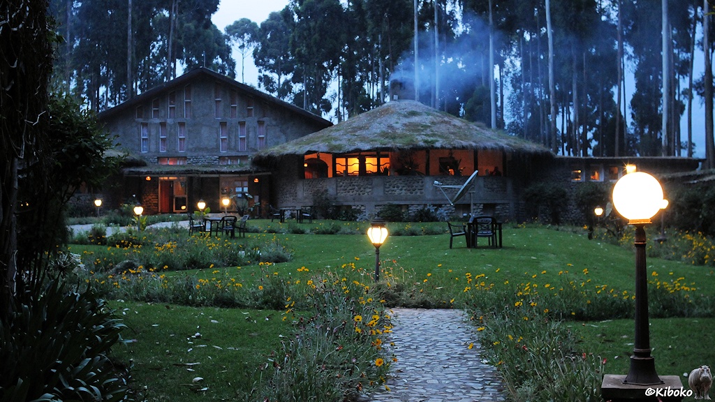 Das Bild zeigt deas Hauptgebäude der Lodge aus grauem Naturstein. Der Schornstein auf dem runden Restaurantanbau stößt blauem Qualm aus. Im Restaurant brennt goldbraunes Licht. Im Garten vor dem Restaurant sehen kleine Laternen.