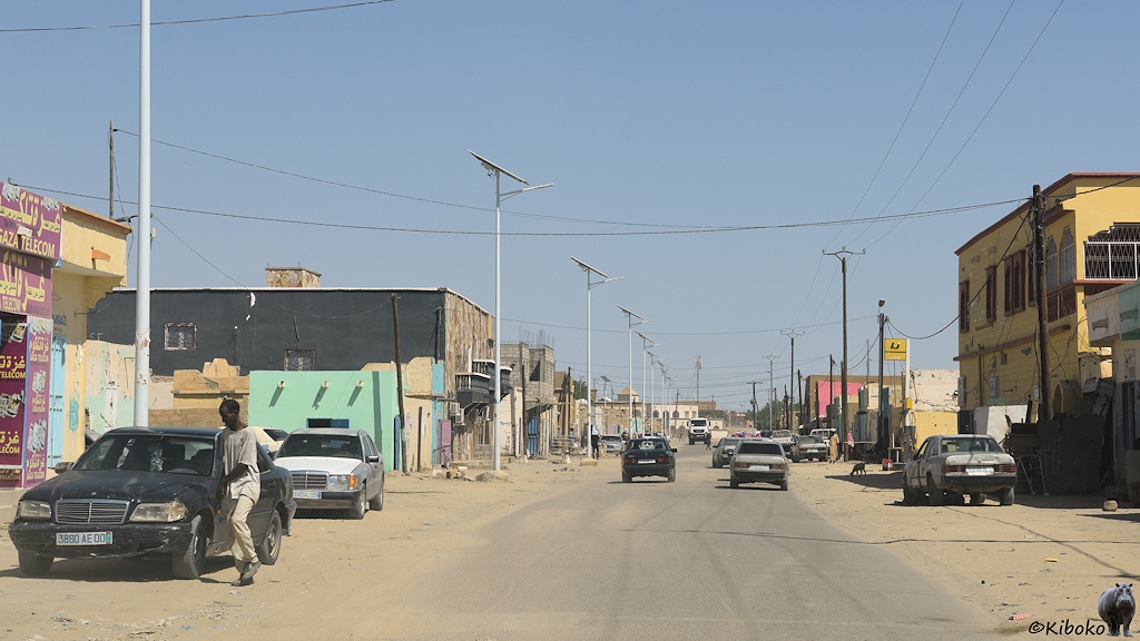 Das Bild zeigt eine staubige Teerstraße. Auf beiden Seiten stehen alte Mercedes Limousinen. Helle, solarbetriebene Straßenlaternen stehen auf der linken Straßenseite. An der Straße stehen einzelne ein- und zweigeschossige Steinhäuser mit Werkstätten und Handwerksbetrieben.