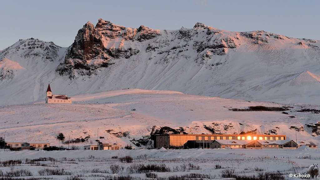 Das Bild zeigt einen Teil einer Ortschaft in einer Winterlandschaft vor einem Gebirgszug im morgendlichen Sonnenlicht. Unten rechts ist ein großes, zweistöckiges, kastenförmiges Gebäude in dessen Fenster sich die Sonne spiegelt. Über den Häusern ist auf einem Hügel eine weiße Kirche mit rotem Dach und viereckigen Turm über dem Eingang.
