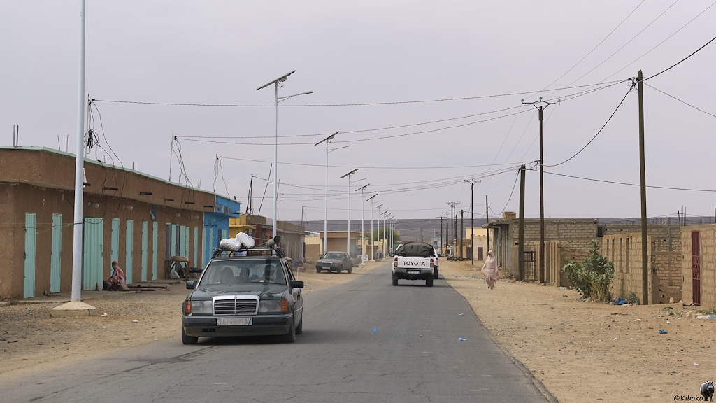 Das Bild zeigt eine gerade Teerstraße. Auf beiden Straßenseiten stehen sandfarbene, einstöckige Gebäude und Mauern. Am linken Straßenrand sind hohe Straßenlaternen mit schrägstehenden Solarpanelen an der Lampenspitze. Ein dunkler Mercedes kommt mit einer Dachladung aus Säcken, Kisten und Rohren entgegen.