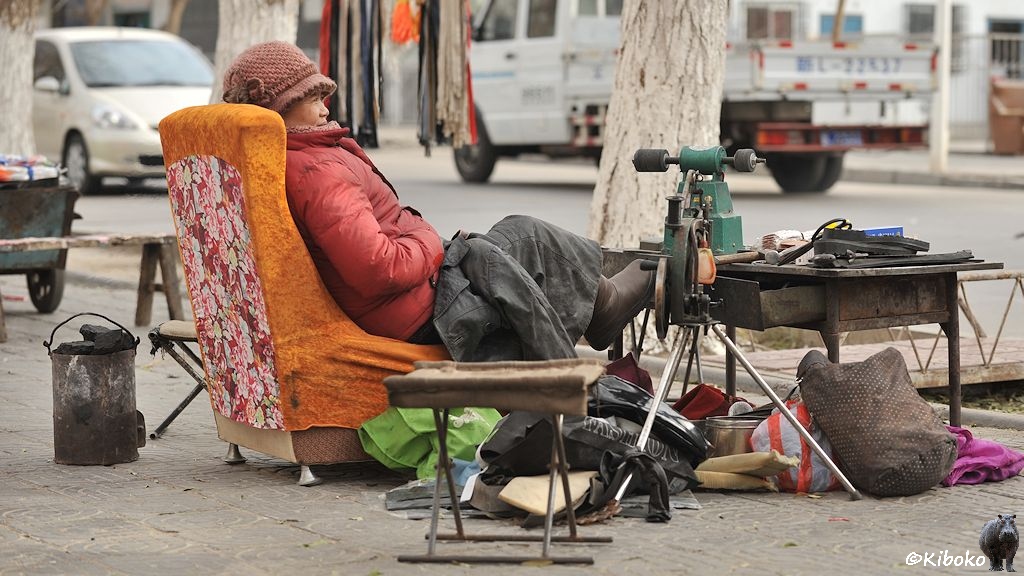 Das Bild zeigt eine ältere Frau in roter Jacke mit Mütze auf einem orangenen Plüschsessel sitzend. Link daneben steht ein Blecheimer mit Kohlen. Rechts steht ein Arbeitstisch aus Blech mit einer Schleifmaschine. Daneben steht eine einfache Nähmaschine auf einem Dreibein. Drumherum stehen Taschen, Beutel und Schuhe.