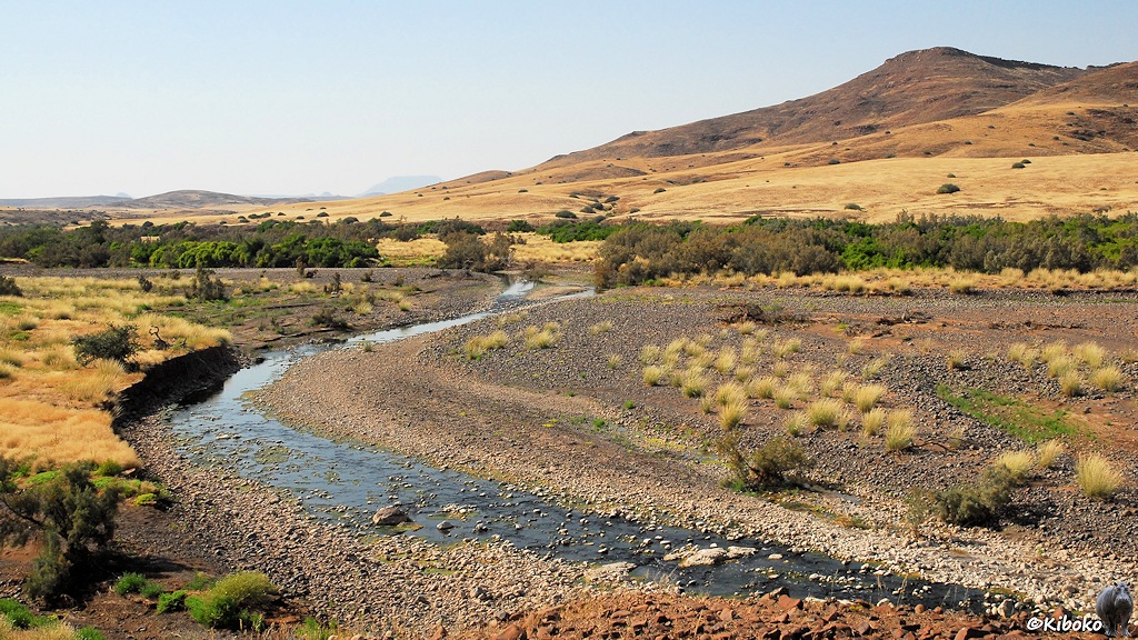 Ein Fluß führt nur wenig Wasser und schlängelt sich über eine Schotterebene. Im Hintergrund sind Büsche im Tal und dahinter Berge.