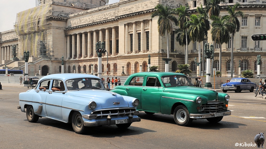 Das Bild zeigt zwei Autos, die nebeneinander vor einem monumentalen Gebäude fahren. Das vordere ist hellblau, das hintere ist grün-metallic lackiert.