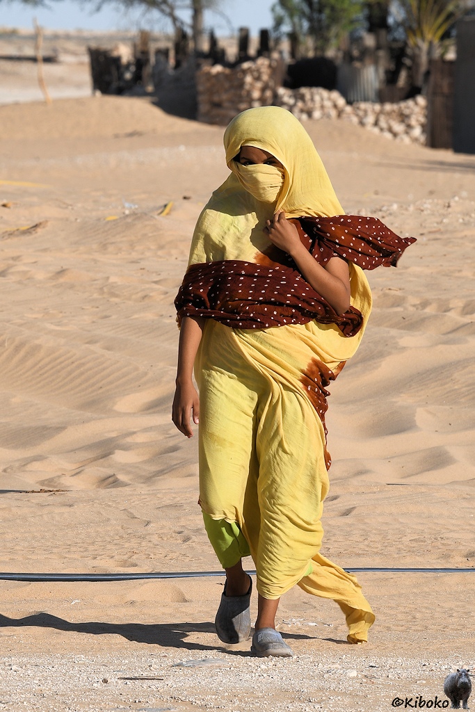 Das Bild zeigt eine Frau im gelben Kleid im Hochformat. Die Haare sind durch ein gelbes Kopftuch verdeckt. Ein gelber Schleier bedeckt das Gesicht. Nur die Augen schauen noch heraus. Um die Arme und Bauch trägt die Frau ein rotbraunes Tuch mit weißen Tupfen.