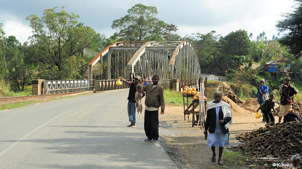 Eine Teestraße und eine Bahnlinie führen über eine Gitterbrücke mit drei Trägern. Die Eisenbahn ist auf der linken Seite. Vor der Brücke steht ein Verkäufer mit Fisch und ein Verkäufer mit Essen in gelben Plastiktüten. Neben der Brücke ist ein Verkaufsstand mit gelben Melonen.