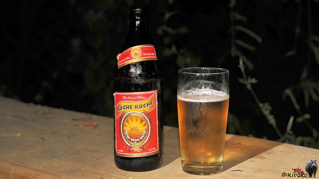Das Bild zeigt eine Bierflasche mit rotem Ettikett mit schwarzer Schrift auf goldenem Grund: Kuche Kuche. Auf einem goldenen Sonnensymbol steht in weißer Schrift Mowa Wathu Wathu. Oben steht auf dem Ettikett: The Pride of Malawi. Danaben steht ein volles Bierglas.