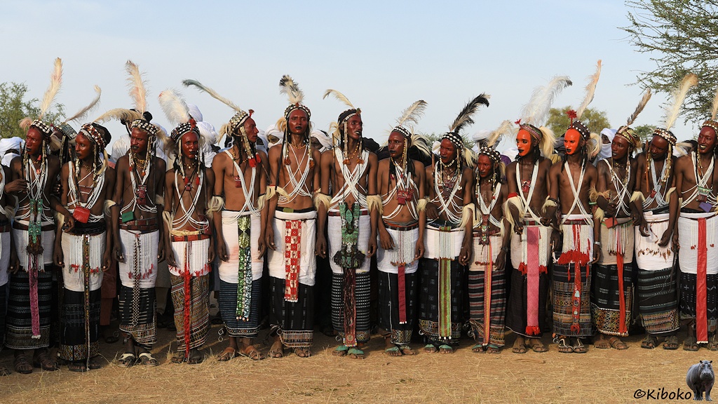 Das Bild zeigt eine Reihe von 15 Männern am Rand der Arena in traditioneller Kleidung. Zwei haben ein orange gefärbtes Gesicht. Die anderen haben ein dunkelrot gefärbtes Gesicht. 12 Männer schauen nach links.
