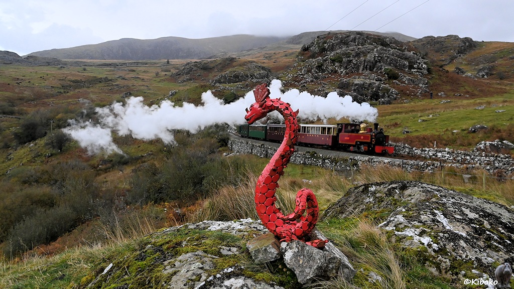 Das Bild zeigt einen roten Metalldrachen der sich s-förmig aufgerichtet hat. Im Hintergrund fährt ein Zug mit einer roten Dampflok und vier Personenwagen vorbei. Im Hintergrund ist ein Bergmassiv aus grauen Felsen.
