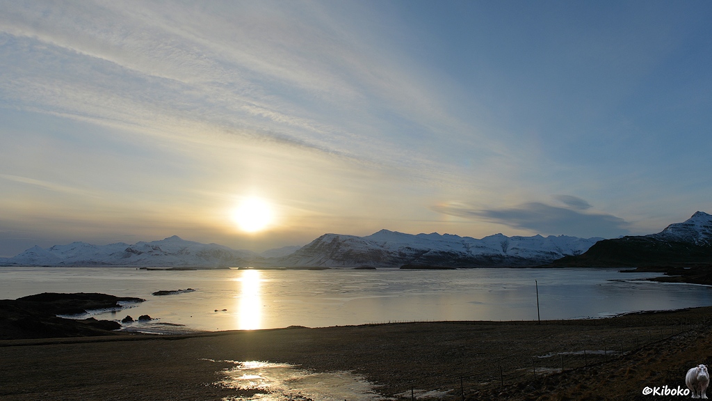 Das Bild zeigt eine weitläufige Landschaftsaufnahme mit einer zugefrorenen Bucht. Die untergehende Sonne spiegelt sich auf dem Eis. In der Ferne ist eine schneebedeckte Bergkette. Am blauen Himmel ziehen diagonal ein paar streifenförmige dünne Wolken durch.