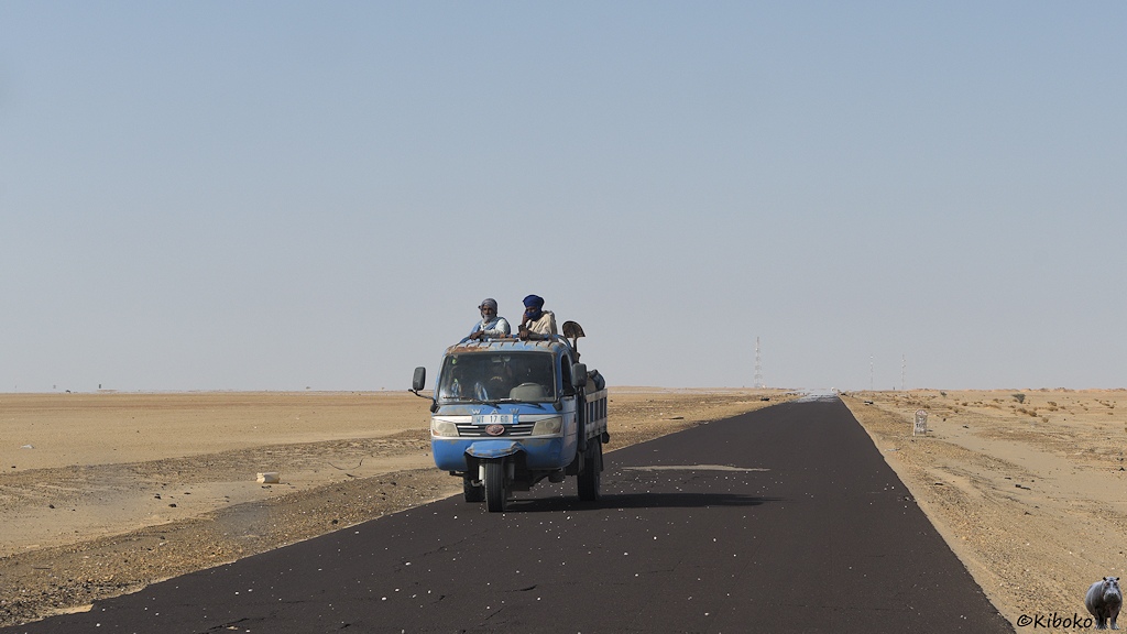 Das Bild zeigt eine Teerstraße die geradeaus ins Bild führt. Um die Straße ist eine hellbraune Schotterwüste. Ein blaues, dreirädriges Fahrzeug mit Pritsche kommt entgegen. Auf der Ladefläche stehen zwei Männer.