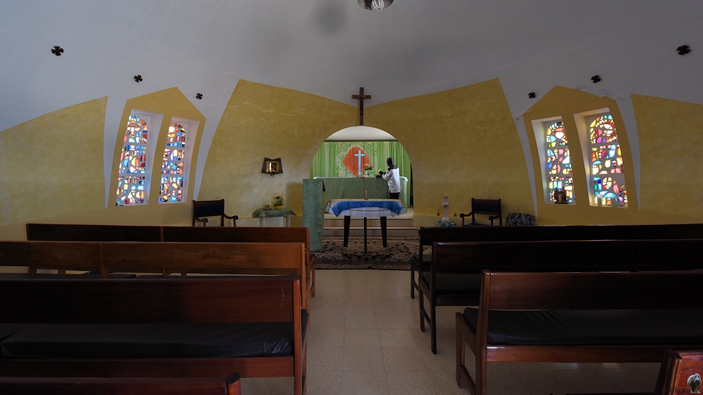 Das Bild zeigt die innenansicht der kugelförmigen Kirche. Auf jeder Seite stehen drei Holzbänke mit jeweils 4 Sitzplätzen. Die Wände sind unten gelb und oben weiß gestrichen. Auf jeder Seite ist ein Doppelfenser mit buntem Glasmosaik. Am Ende der Kirche ist ein runder Durchlass in den Altarraum. Dort steht ein schlichtes weißes Kreuz vor einem grünen Vorhang mit einem orangeroten Fleck hinter dem Kreuz. Ein Pastor in weißem Hemd steht vor dem Altar.
