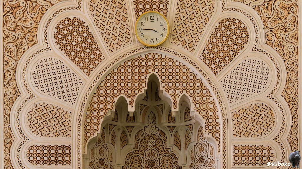Das Bild zeigt den Oberteil einer Nische, die durch einen Bogen mit 9 Blättern umgeben ist. Die Blätter sind mit unterschiedlichen, filigranen Steingravuren geschmückt. Darüber hängt eine runde Uhr mit Goldrand und der Aufschrift: Quarz.