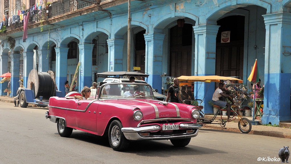 Das Bild zeigt ein altes rosa Cabrio amerikanischer Bauart. Das Verdeck ist offen. Eine Frau sitzt auf dem Rücksitz. Das Auto hat zwei breite Zierleisten in Längsrichtung auf der Motorhaube. Im Hintergrund ist ein hellbrauer Arkardengang mit viereckgien Säulen. Die untere Hälfte der Säulen ist himmelblau. Im Hinergrund wartet eine Fahrradrikscha. Hinter dem Auto steht ein einachsiger Anhänger mit einer großen Kabeltrommel.