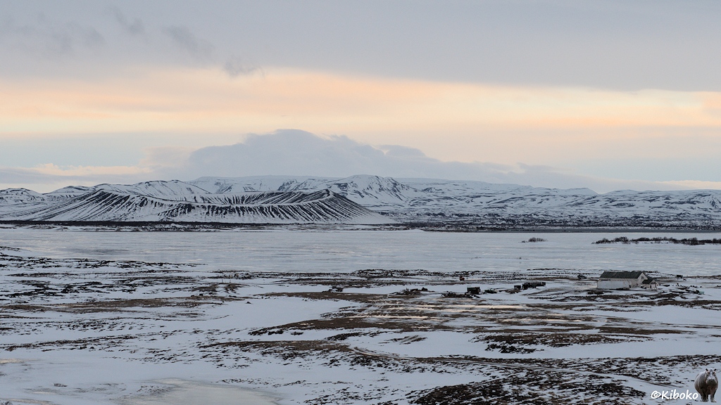 Landschaftsaufnahme mit Blick über ein breites, schneebedecktes Tal. Im Zentrum ist ein großer See, der mit Eisschollen bedeckt ist. Hinter dem See ist ein Vulkankrater.