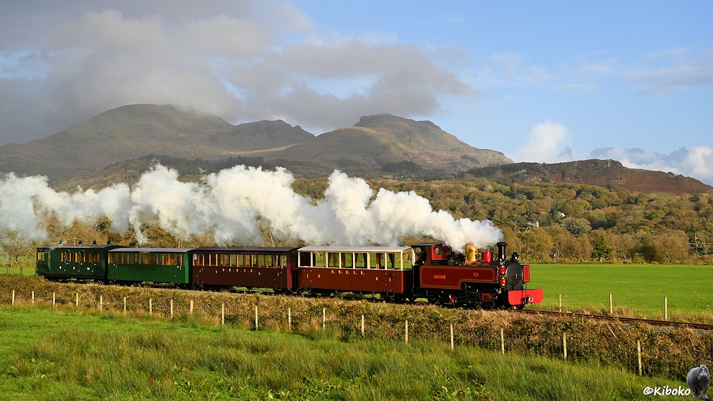 Das Bild zeigt eine rote Tenderlokomotive mit schwarzem Kessel und goldenem Dom mit vier Personenwagen über eine Wiesenlandschaft fahren. Die Lok zieht vier Personenwagen und hat eine lange weiße Dampffahne. Im Hintergrund sind hohe, baumlose Berge. Der höchste Gipfel ist von Wolken verhüllt.
