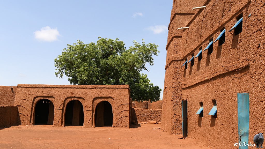 Das Bild zeigt die Fassade der Moschee am rechten Bildrand. Link im Hintergrund steht eine offene Halle aus Lehm mit drei Bögen. Davor ist ein Platz aus rötlichem Sand.