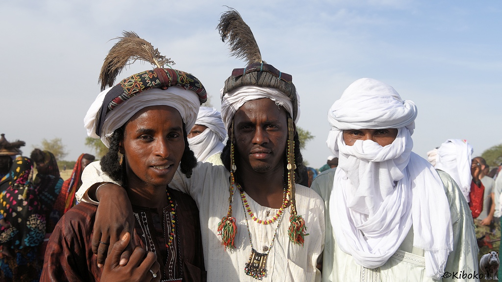 Das Bild zeigt das Porträt von drei Männern. Die linken beiden tragen eine gewickelte Kopfbedeckung mit Feder und Halsschmuck. Der rechte Mann hat sich mit einem weißen Turban das Gesicht verdeckt. Nur die Augen schauen noch raus.