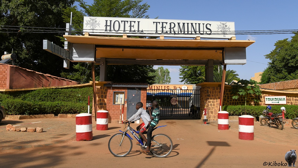 Das Bild zeigt die Eingangspforte zum Hotel Terminus. Die Einfahrt ist durch rot-weiß-rote Tonnen gesichert. Über der Pforte ist ein Dach mit der Aufschrift: Hotel Terminus. Im Vordergrund fahrn zwei Kinder auf einem Fahrrad vorbei. Mangels Gepäckträger quetschen sich beide auf den Sattel.