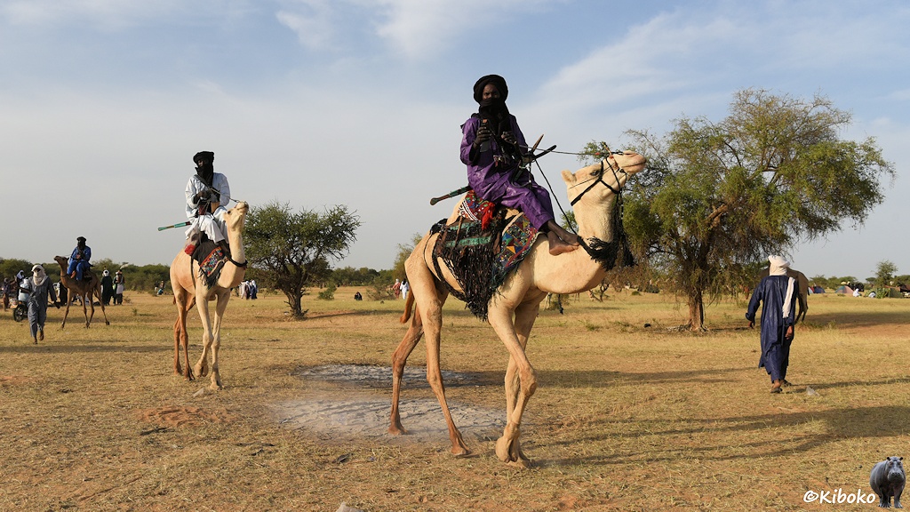 Das Bild zeigt zwei Männer auf Dromedaren reitend. Der erste Mann trägt ein violettes Gewand und schwarzem Turban. Der hintere Mann trägt ein weißes Gewand und ebenfalls einen schwarzen Turban. Im Hintergrund ist eine Savannenlandschaft mit einzelnen größeren Akazienbäumen.