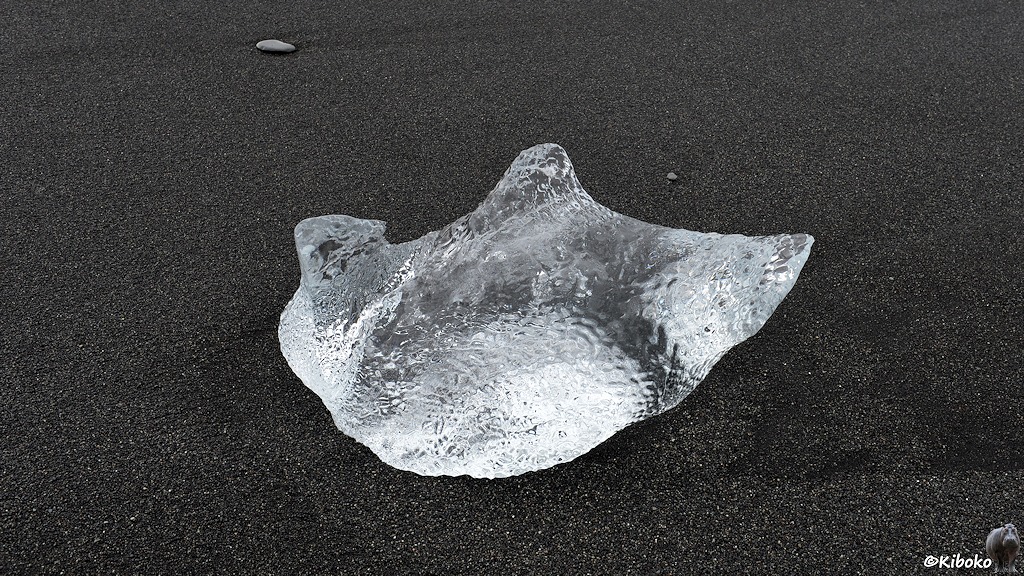 Das Bild zeigt einen kleineren Eisblock mit drei abgerundet spitzen auf schwarzem Sand. Auch dieser Eisblock hat wieder eine Dellentextur.
