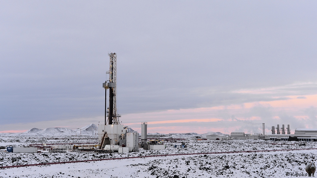 Das Bild zeigt eine schneebedeckte Felslandschaft. Darin steht ein Turm aus einem Gittermast. Im Hintergrund sind weitere Gebäude und Gittertürme. Aus denen steigt Dampf in den Himmel.
