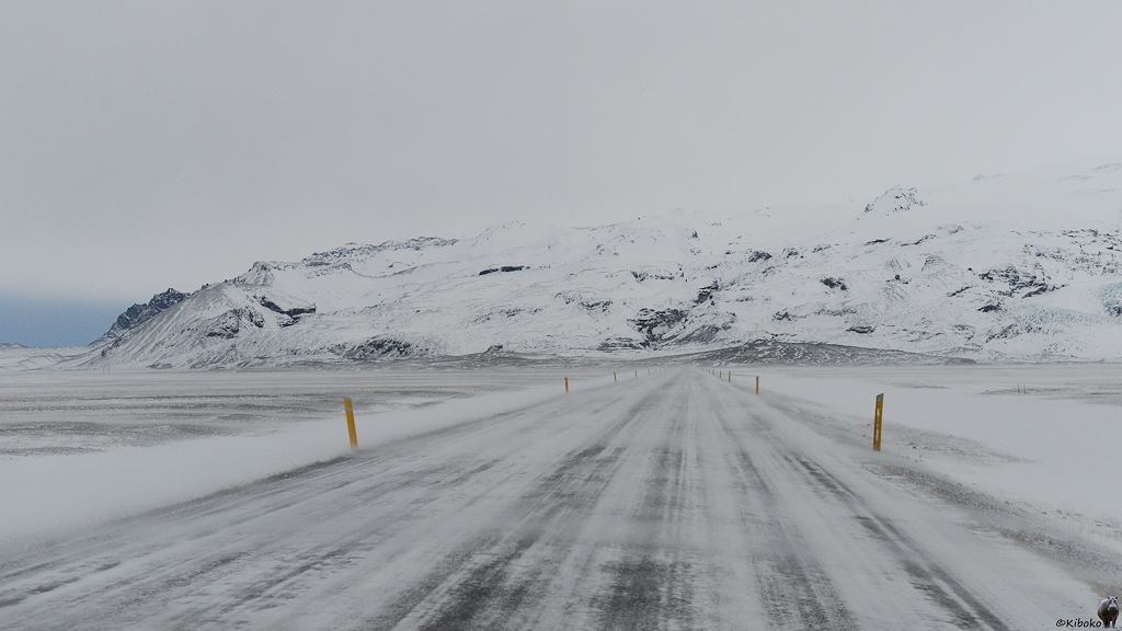 Das Bild zeigt eine vereiste Straße die gerade auf ein Gebirgszug führt. Gelbe Leitpfähle begrenzen die Fahrbahn. Der Wind weht Schnee über die Straße.