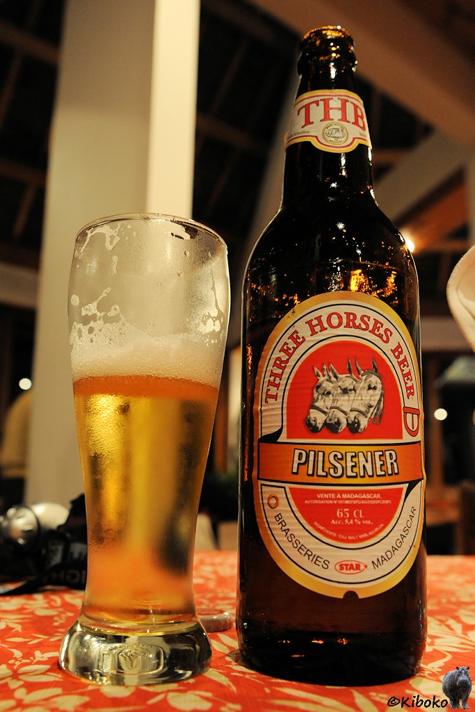 Gefüllte Bierglas und Bierflasche mit rotem Etikett auf dem drei Pferdeköpfe abgebildet sind.
