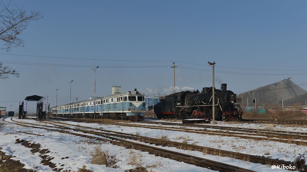 Das Bild zeigt eine Reihe von sechs weiß-blauen Diesellokomoitven auf dem Abstellgleis. Links daneben fährt eine Dampflok vorbei. Im Hintergrund sind Abraumhalden.