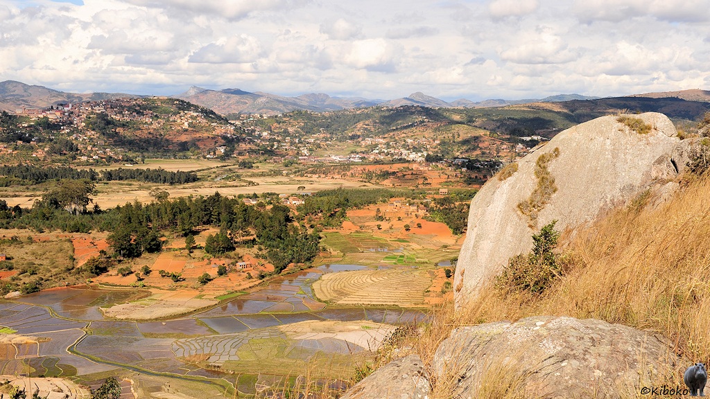 Das Bild zeigt einen Blick auf einen entfernte Hügelkette mit der Stadt Fianarantsoa. Im Fordergrund ist ein großer grauer Fels und trockenes Gras. Im Tal sind geflutete Reisfelder und trockene Felder.