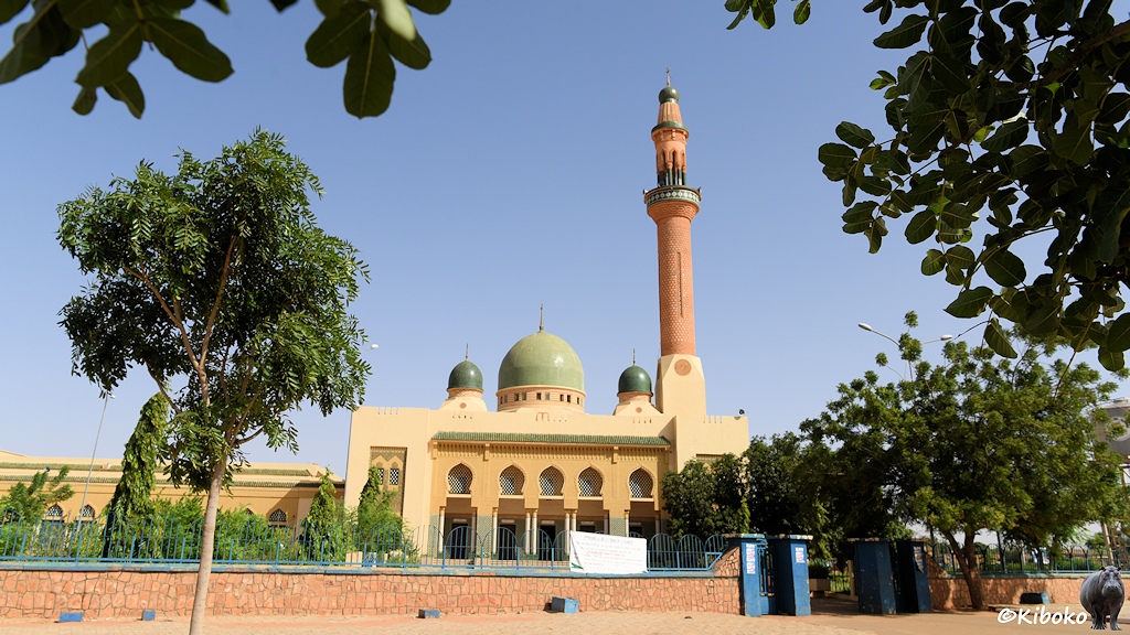 Das Bild zeigt eine Moschee aus beigen Steinen mit drei grünen Kuppeln und einem hohen rötlichen Minarett. Das Bild wurde aus einem Park aufgenommen und runherum ragen Bäume und Blätter ins Bild.