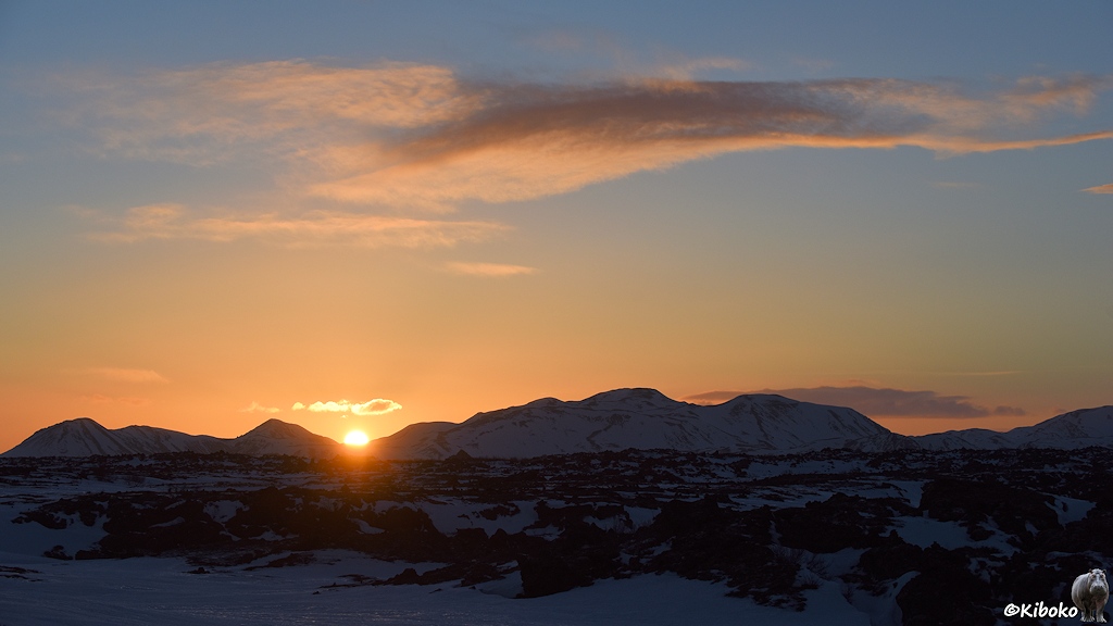 Die Sonne geht in einer Talsenke einer Bergkette auf. Der Himmel leuchtet in Orange. Im Vordergrund sind Lavafelder mit Schneefeldern