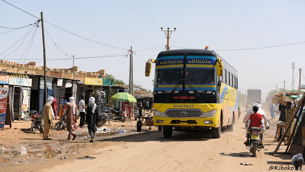 Das Bild zeigt einen gelben Reisebusauf einer sandigen Straße. In die Gegenrichtung fährt ein Motorrad. Auf beiden Seiten der Straße sind einstöckige Geschäfte. Am Straßenrand sind Pfützen, in denen Müll schwimmt.