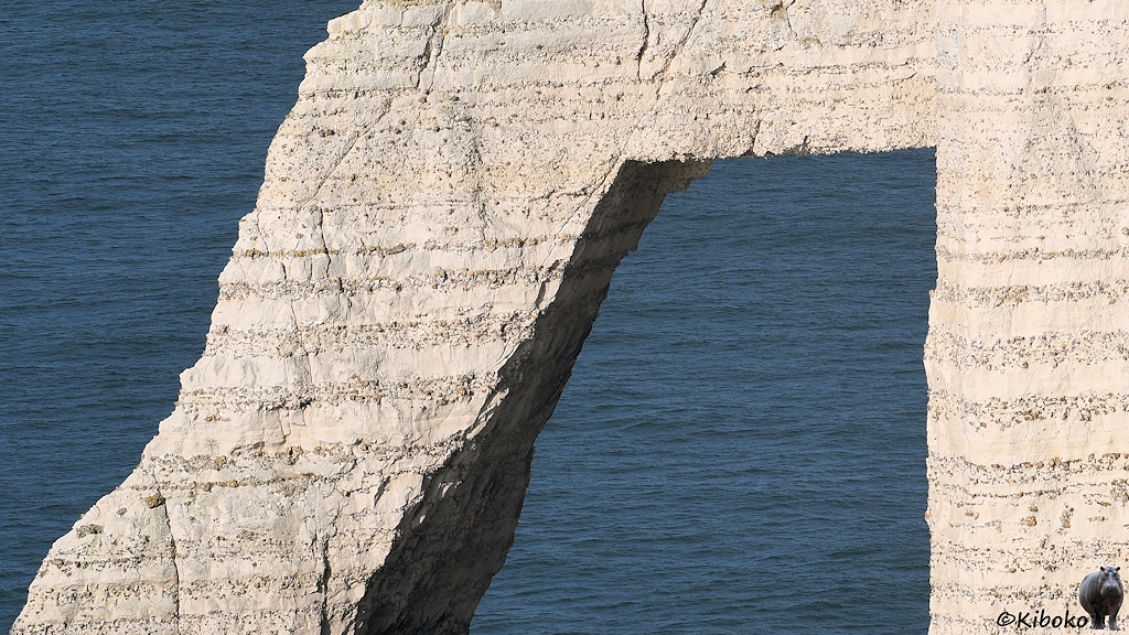 Das Bild zeigt einen kleinen Ausschnitt eines Bogens an der Steilküste. Es zeigt den Schichtaufbau aus Lagen mit feinkörnigen Material und Lagen mit eingelagerten Steinen. Der Bogen hat eine geraden Abschluss entlang einer Lage. Die Linke Seite ist ein schmaler Pfeiler. Dahinter ist das dunkelblaue Meer.