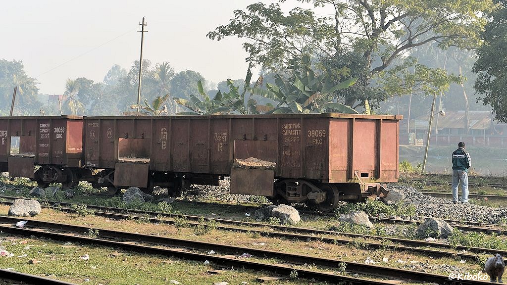 Das Bild zeigt einen braunen vierachsigen offenen Güterwagen mit zwei Klappen auf jeder Seite. Die Seitenklappen sind offen. Im Wagen liegen nur noch Reste der Ladung. Hinter dem Wagen liegt ein Haufen hellgrauer Schotter. Vor dem Wagen liegen einzelne graue Felsbrocken.