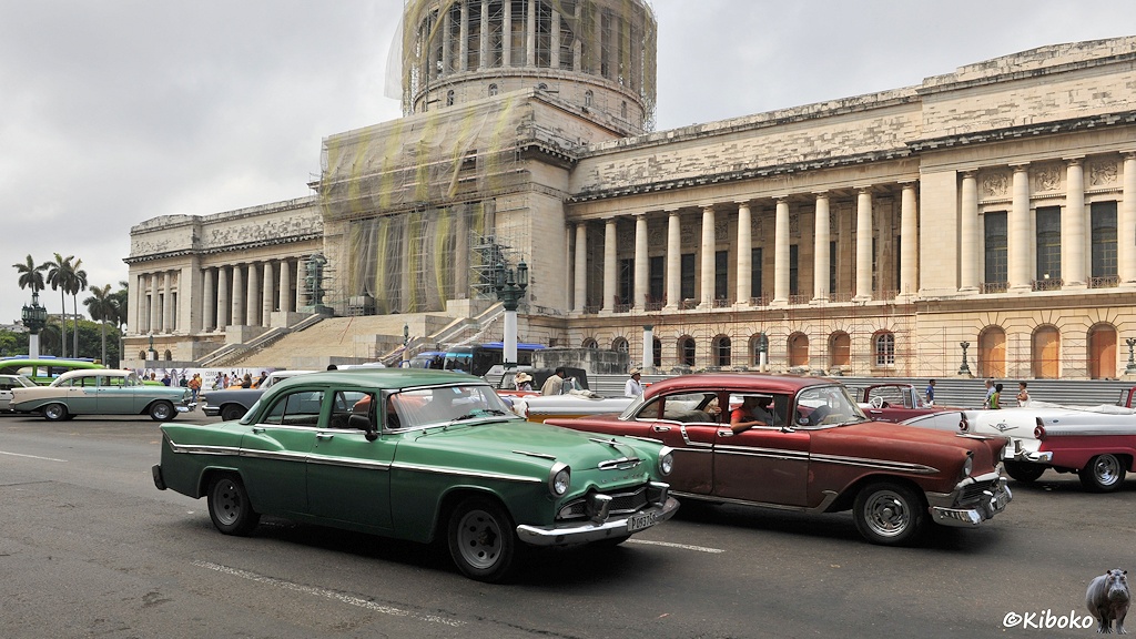 Das Bild zeigt zwei Autos in grün und rot vor einem monumentalen Gebäude. Das Gebäude hat eine Fassade mit hohen Säulen. Der Mittelteil des Gebäudes ist mit Netzen behangen. Darüber ist eine runde, mit Säulen umgebene Kuppel, die ebenfalls mit Nezten verhüllt ist.