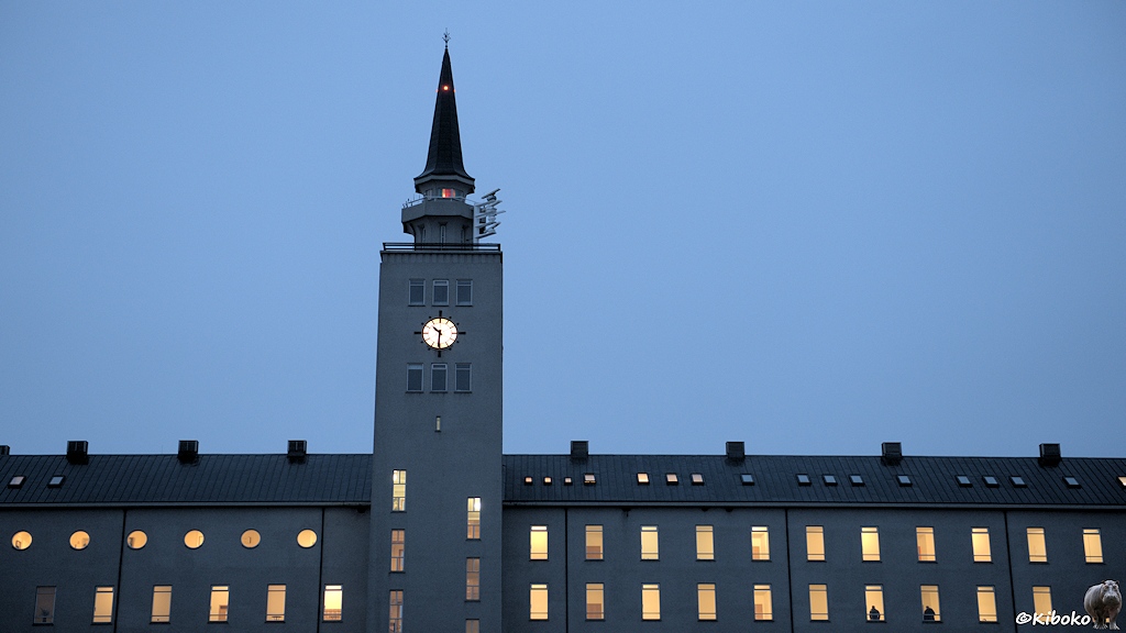 Während der Dämmerung sind bei einem mehrstöckigem Gebäude alle Fenster hell erleuchtet. Ein viereckiger Turm mit langer Turmspitze lockert die Fassade auf. Die beleuchtete Turmuhr zeigt 10:30 Uhr.