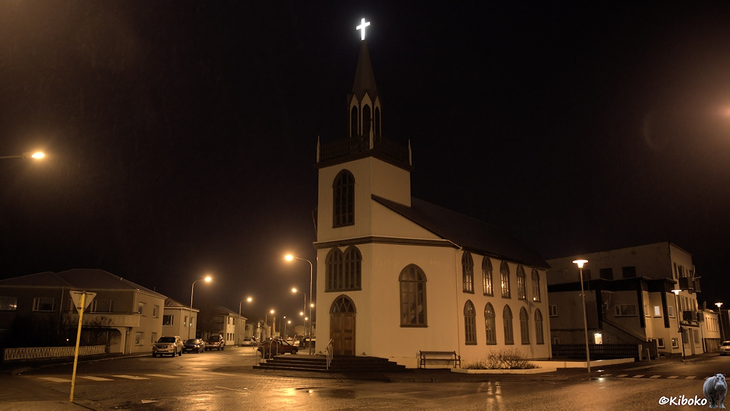 An einer nächtlichen Straßenkreuzung steht eine weiße Kirche mit einem spitzen Turm über der Eingangstür. Auf der Turmspitze ist ein weißes, beleuchtetes Kreutz