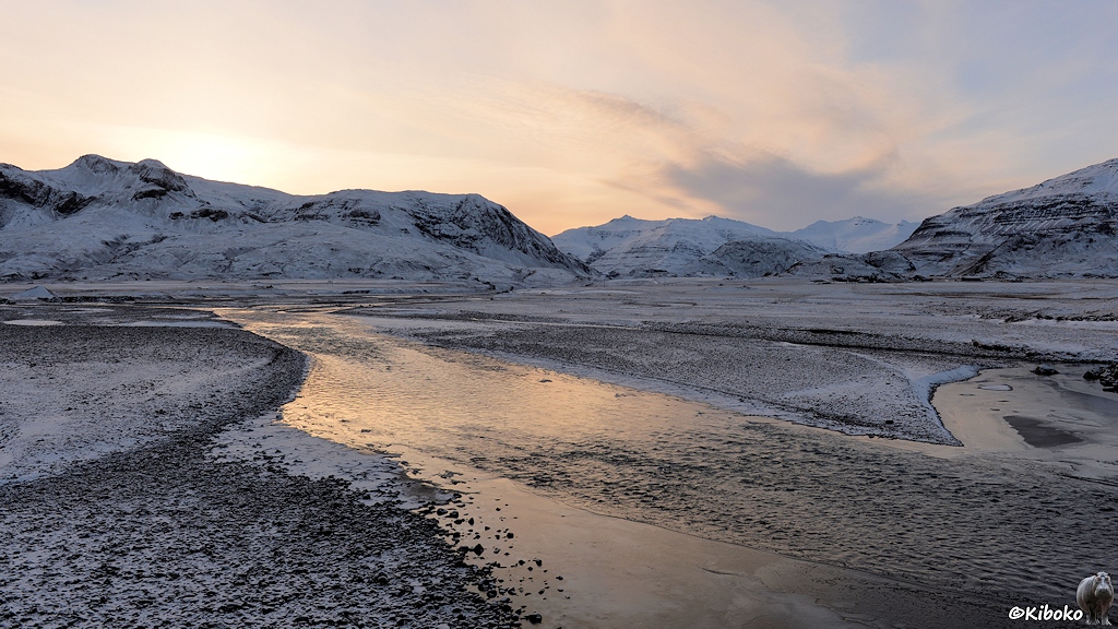 Das Bild zeigt eine schwarze Schotterebene mit einer ganz dünnen Schneeschicht. Dazwischen sind verzweigte Arme eines flachen Flusses in denen sich das Licht spiegelt. Im Hintergrund sind schneebedeckte Berge, hinter denen die Sonne gerade versunken ist. Der Himmel leuchtet in sehr blassen Orangetönen.