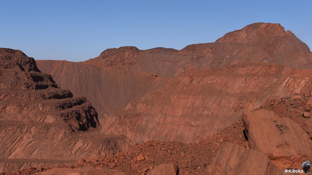Blick in die Eisenmine aus rotem Gestein. Ein LKW verlässt als winziger gelber Punkt die Mine am gegenübeliegenden Berghang.