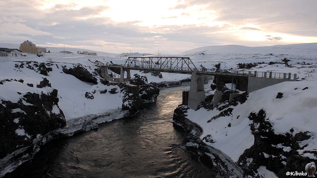 Eine Gitterbrücke mit Betonpfeilern spannt sich über eine Schlucht mit einem Fluss. Am linken Bildrand steht ein größeres, gelbes Holzgebäude