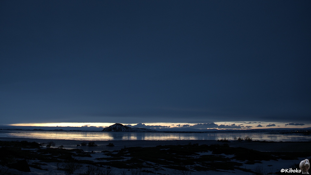 Landschaftsaufnahme unter einen dunklen blau-grauen Himmel. Ein schmaler heller Lichtstreifen am Horizont spiegelt sich auf dem Eis eines Sees.